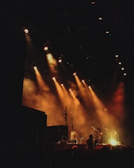 A dimly lit stage with warm, atmospheric lighting casting beams across the scene. Several microphones and musical equipment, including amplifiers and speakers, are visible. The stage is set for a performance with a smoky ambiance lingering in the air. In the foreground, a person is capturing the scene with a camera.