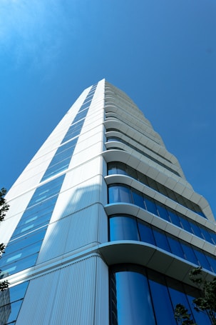 A modern skyscraper with a sleek, white facade contrasted by reflective blue-tinted windows. The structure has a series of curved balconies or ledges extending outwards as it rises into a clear blue sky. The image captures the building from a low angle, emphasizing its height and architectural elegance.