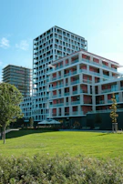 Modern multi-story buildings with geometric designs are surrounded by greenery, including a lush lawn and trees. The structures have a mix of red and white colors with extensive use of glass, creating a clean and contemporary aesthetic.