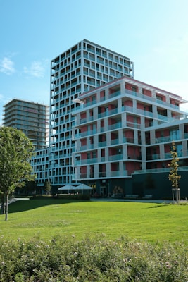 Modern multi-story buildings with geometric designs are surrounded by greenery, including a lush lawn and trees. The structures have a mix of red and white colors with extensive use of glass, creating a clean and contemporary aesthetic.