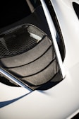 A detailed view of a car's air intake or vent area, featuring sleek carbon fiber textures and a modern design. The surface reflects light, highlighting the geometric patterns. The surrounding area is smooth, possibly indicative of a sports car body.