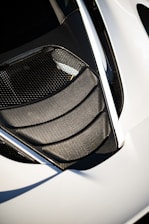 A detailed view of a car's air intake or vent area, featuring sleek carbon fiber textures and a modern design. The surface reflects light, highlighting the geometric patterns. The surrounding area is smooth, possibly indicative of a sports car body.