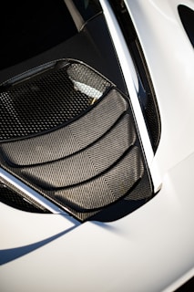 A detailed view of a car's air intake or vent area, featuring sleek carbon fiber textures and a modern design. The surface reflects light, highlighting the geometric patterns. The surrounding area is smooth, possibly indicative of a sports car body.