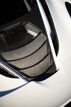 A detailed view of a car's air intake or vent area, featuring sleek carbon fiber textures and a modern design. The surface reflects light, highlighting the geometric patterns. The surrounding area is smooth, possibly indicative of a sports car body.
