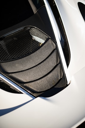 A detailed view of a car's air intake or vent area, featuring sleek carbon fiber textures and a modern design. The surface reflects light, highlighting the geometric patterns. The surrounding area is smooth, possibly indicative of a sports car body.