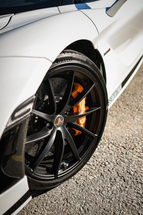 A close-up view of a car's black alloy wheel with orange brake calipers against a smooth gravel background. The car features a sleek, aerodynamic design with a glossy white paint finish.