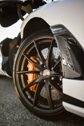 A close-up view of a high-performance car's front wheel, featuring a sleek black alloy rim with an intricate design. The bright orange brake caliper, marked with the word 'MCLAREN,' is visible through the spokes. A section of the white car body, including aerodynamic components, is partially visible. The surface below is textured asphalt, suggesting a road or track setting.