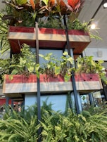 A vertically arranged, multi-level planter structure with lush greenery. The planters are wooden with red and natural wood finish, supported by black metal frames. Various types of plants, including ferns and other leafy vegetation, are growing at different levels. Natural light illuminates the area, giving it a fresh and vibrant atmosphere. In the background, a glimpse of an indoor setting with ceiling lights and part of a building interior is visible.