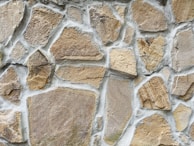 A close-up view of a stone wall with irregularly shaped stones set in light gray mortar. The stones vary in size and texture, featuring shades of beige, tan, and light brown.