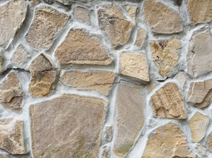 A close-up view of a stone wall with irregularly shaped stones set in light gray mortar. The stones vary in size and texture, featuring shades of beige, tan, and light brown.