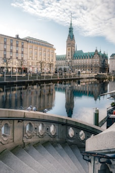 A scenic European cityscape featuring an elegant staircase in the foreground, leading down to a waterfront. The backdrop showcases a historic building with a tall clock tower, with a reflection visible in the calm water. The sky is partly cloudy, adding a peaceful ambiance.