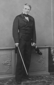 An older man with light-colored hair stands against an ornate backdrop holding a violin and bow. He wears a formal double-breasted coat and matching trousers, complemented by a high-collared shirt. The setting has a classical feel with decorative paneling and a richly patterned carpet.