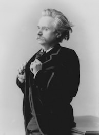 A person with a mustache and styled hair is adjusting their jacket. The image is in black and white, with the person looking off to the side.