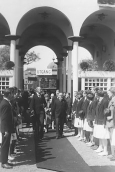 An event with people dressed formally, likely indicative of a ceremonial or official gathering. They stand on either side of a carpet, under large arches with columns, in an outdoor setting. A prominent sign reads 'UTGANG', meaning exit in some Scandinavian languages.
