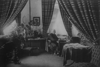 A man sits at a desk in a richly decorated room, surrounded by heavy drapery and various furnishings. Papers and books are spread across the desk and nearby table. There are ornate curtains, plants, and artwork on the walls, creating a classic and intellectual atmosphere.