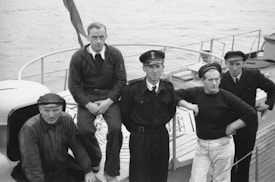 Five men in naval uniforms and casual attire stand and sit on the deck of a ship, with one man leaning against a handrail. The background includes parts of the ship and a visible body of water.