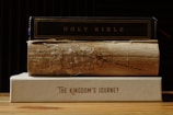 A stack of three books is placed on a wooden surface. The top book is titled 'HOLY BIBLE' with a dark cover. The middle book appears old and worn, with noticeable damage and frayed edges. The bottom book has a clean beige cover and is titled 'THE KINGDOM'S JOURNEY'.