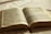 An open book featuring text in a language that appears to be Portuguese. The pages are aged, with dark print and some sections highlighted in pink and blue. A red bookmark peeks out from the bottom, and the book is lying on a white fabric surface with soft lighting, creating a warm and serene atmosphere.