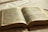 An open book featuring text in a language that appears to be Portuguese. The pages are aged, with dark print and some sections highlighted in pink and blue. A red bookmark peeks out from the bottom, and the book is lying on a white fabric surface with soft lighting, creating a warm and serene atmosphere.