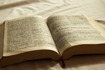 An open book featuring text in a language that appears to be Portuguese. The pages are aged, with dark print and some sections highlighted in pink and blue. A red bookmark peeks out from the bottom, and the book is lying on a white fabric surface with soft lighting, creating a warm and serene atmosphere.