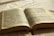 An open book featuring text in a language that appears to be Portuguese. The pages are aged, with dark print and some sections highlighted in pink and blue. A red bookmark peeks out from the bottom, and the book is lying on a white fabric surface with soft lighting, creating a warm and serene atmosphere.
