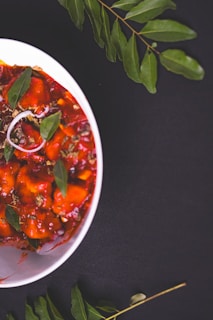 A white bowl contains a vibrant red dish with pieces of food garnished with green leaves, possibly curry leaves. The dish has a rich, saucy texture with visible herbs and spices. Surrounding the bowl are curry leaves scattered on a dark surface, creating a striking color contrast.