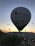 A large hot air balloon is prominently in the foreground, with several other smaller balloons scattered across the sky as the sun rises or sets on the horizon. The landscape below shows hills and sparse vegetation, while the balloon displays a logo and website. The scene conveys a sense of adventure and serenity.