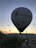 A large hot air balloon is prominently in the foreground, with several other smaller balloons scattered across the sky as the sun rises or sets on the horizon. The landscape below shows hills and sparse vegetation, while the balloon displays a logo and website. The scene conveys a sense of adventure and serenity.