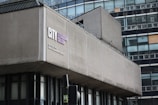 A modern, urban building with a facade marked by rectangular windows and concrete panels. The sign reads City of Glasgow College situated on City Campus, North Hanover Street. A traffic light and part of another structure are visible in the foreground.