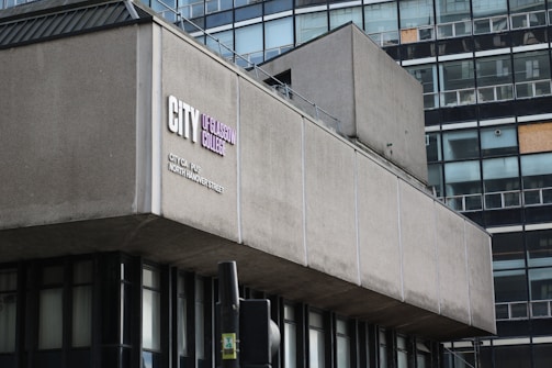 A modern, urban building with a facade marked by rectangular windows and concrete panels. The sign reads City of Glasgow College situated on City Campus, North Hanover Street. A traffic light and part of another structure are visible in the foreground.