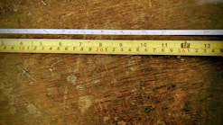 Two tape measures are placed parallel on a wooden surface. The upper tape measure is white with black and red markings, while the lower one is yellow with similar markings. The wooden surface shows signs of wear and has an uneven texture.