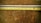 Two tape measures are placed parallel on a wooden surface. The upper tape measure is white with black and red markings, while the lower one is yellow with similar markings. The wooden surface shows signs of wear and has an uneven texture.