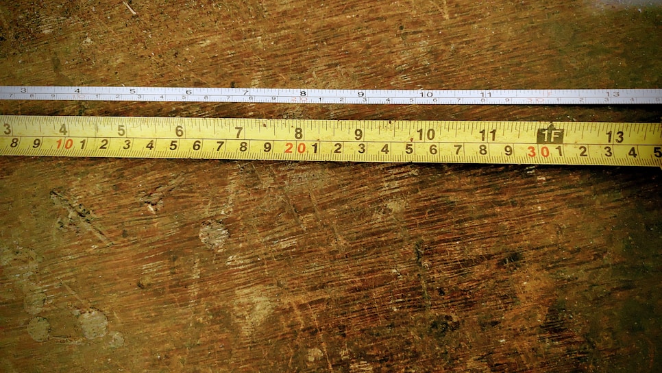 Two tape measures are placed parallel on a wooden surface. The upper tape measure is white with black and red markings, while the lower one is yellow with similar markings. The wooden surface shows signs of wear and has an uneven texture.
