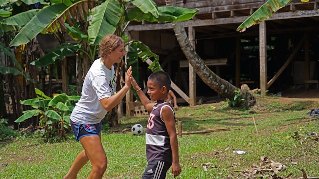 A young woman and a boy enthusiastically engage in a high-five outdoors. They are in a grassy area surrounded by banana plants and a rustic wooden structure in the background. A soccer ball is visible on the ground nearby, indicating play and recreation.
