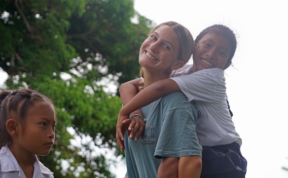 A joyful scene featuring three people in a natural outdoor setting. One person carries another on their back, both smiling and expressing happiness. The third person, standing nearby, looks off to the side with a neutral expression. Lush green trees fill the background.