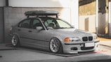 A silver BMW sedan is parked in an indoor area with concrete walls. The car is equipped with a roof box and has aftermarket wheels with a low profile. The lighting creates a soft shadow on the vehicle, highlighting its contours.