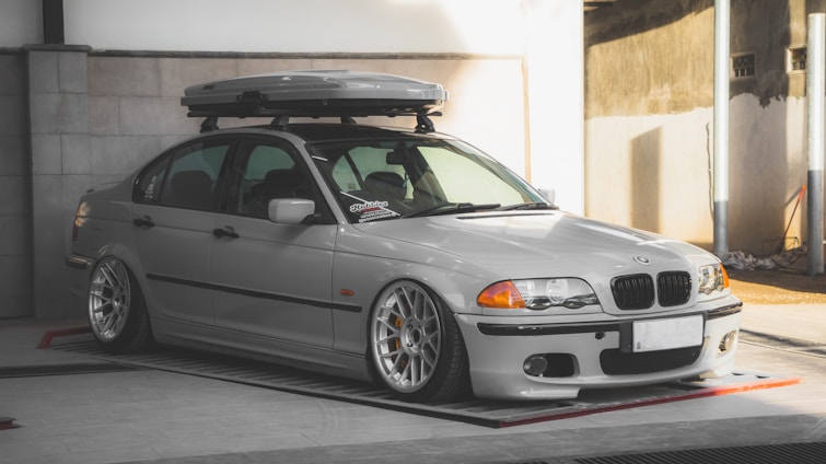 A silver BMW sedan is parked in an indoor area with concrete walls. The car is equipped with a roof box and has aftermarket wheels with a low profile. The lighting creates a soft shadow on the vehicle, highlighting its contours.