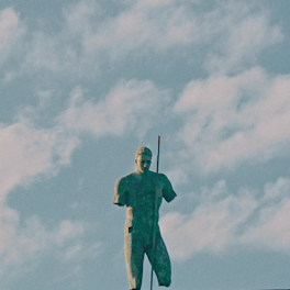 A weathered bronze statue of a muscular male figure holding a long staff, standing against a backdrop of a cloudy sky. Parts of the statue appear eroded, enhancing its antique and stoic presence.