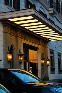 The entrance of a grand building with classic architecture featuring ornate lanterns mounted on stone columns. Large awning with glowing yellow lights and a sign reading 'Hassler Villa Medici'. A person in uniform stands by the door, and a black car is parked in front.