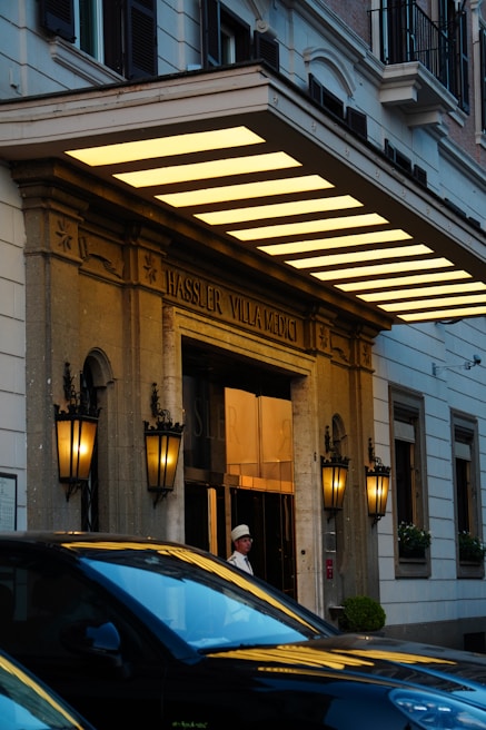 The entrance of a grand building with classic architecture featuring ornate lanterns mounted on stone columns. Large awning with glowing yellow lights and a sign reading 'Hassler Villa Medici'. A person in uniform stands by the door, and a black car is parked in front.