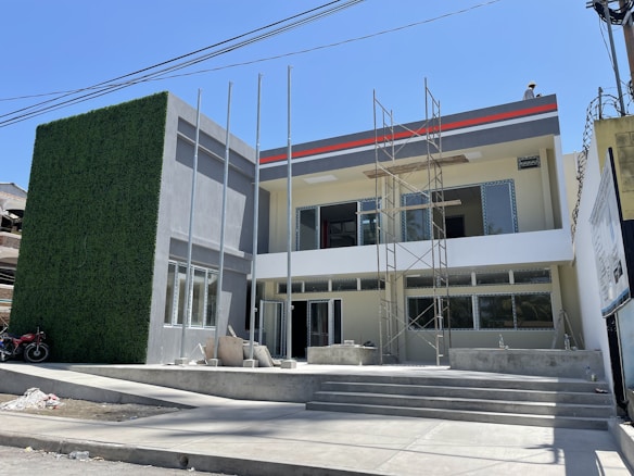 A modern two-story building under construction features a facade with large windows and a decorative wall covered in artificial greenery. Scaffolding is present in front of the building, indicating ongoing work. The structure has a clean, minimalist design with a combination of light gray and white tones.