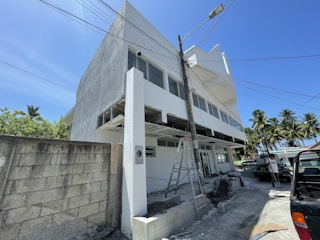 A modern white building is under construction with a visible ladder leaning against it amid construction materials scattered around. Palm trees are present in the background, and several vehicles are parked near the site. The sky is clear and blue, suggesting a sunny day.