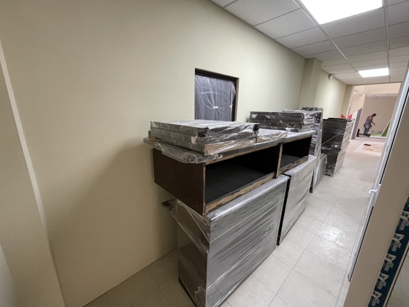 A hallway with beige walls and a tiled floor. Several pieces of furniture covered in plastic wrap are stacked against the wall. At the far end of the hallway, a person is seen working on something bent over.