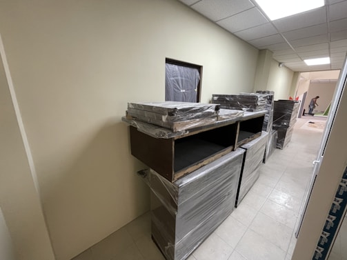 A hallway with beige walls and a tiled floor. Several pieces of furniture covered in plastic wrap are stacked against the wall. At the far end of the hallway, a person is seen working on something bent over.
