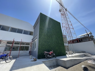 A construction site with a partially finished building, featuring a tall wall covered with green foliage. To the left, there are metal scaffolds and some unfinished sections. A red and white crane towers overhead. Two motorcycles are parked in the foreground, and a dog rests on the ground.