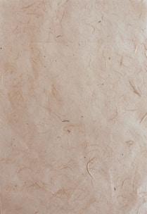 A textured paper surface with a natural, slightly rugged appearance featuring subtle lines and small spots. The paper has a light brown, almost beige hue with visible fibers embedded within the material.