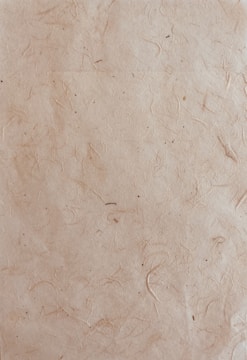 A textured paper surface with a natural, slightly rugged appearance featuring subtle lines and small spots. The paper has a light brown, almost beige hue with visible fibers embedded within the material.