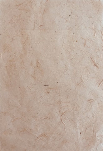 A textured paper surface with a natural, slightly rugged appearance featuring subtle lines and small spots. The paper has a light brown, almost beige hue with visible fibers embedded within the material.