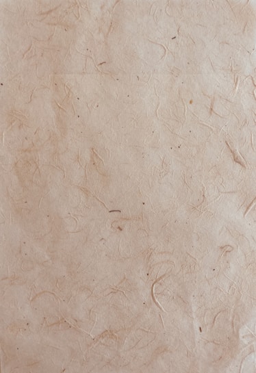 A textured paper surface with a natural, slightly rugged appearance featuring subtle lines and small spots. The paper has a light brown, almost beige hue with visible fibers embedded within the material.