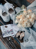 A wedding-themed arrangement with a pair of white sneakers adorned with lace, a bouquet of cream-colored roses with baby's breath, elegant invitations with a floral design and wax seal, two matching wristwatches, a pair of wedding rings, and delicate jewelry all placed on rustic wooden planks. Translucent white tulle drapes gracefully over several of these items, adding a touch of sophistication.